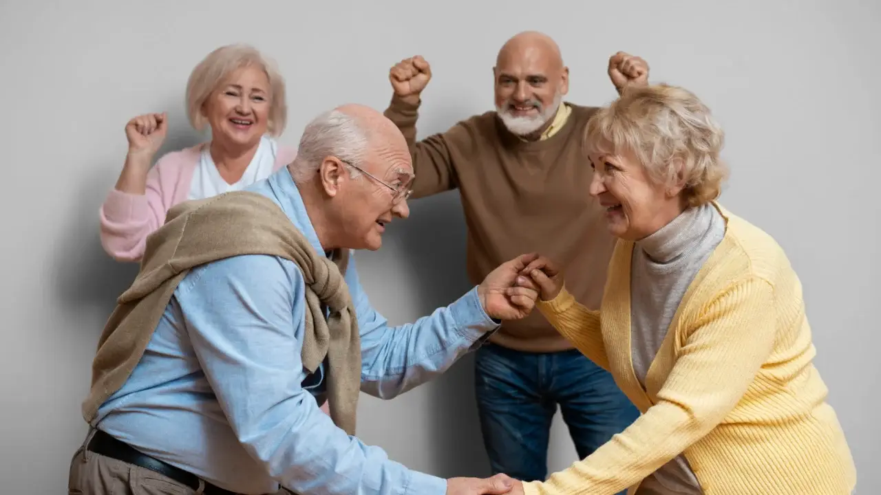 Fyra leende äldre vuxna står inomhus; två framför dansar medan de andra två hejar på dem i bakgrunden. Alla verkar glada och är klädda i avslappnade, färgglada tröjor. Bakgrunden är enfärgad och ljus.