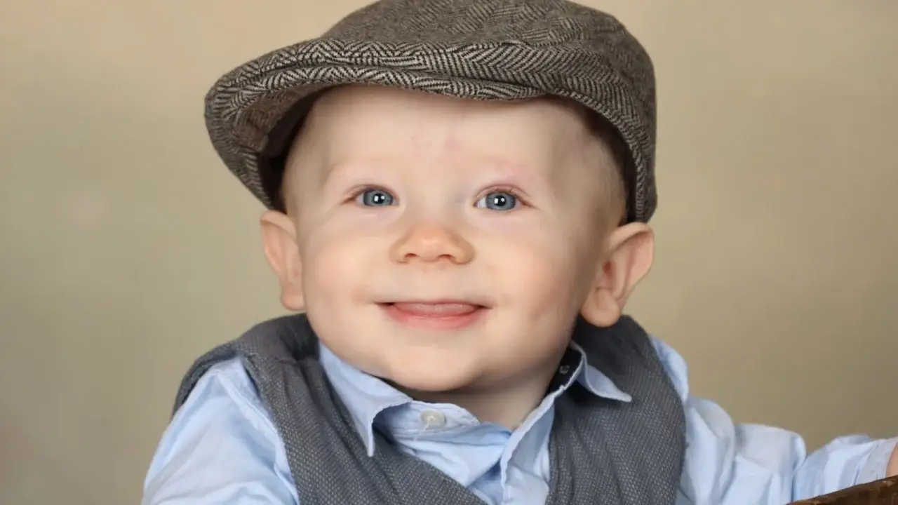 En leende baby iförd en grå flat keps, en ljusblå skjorta och en grå väst tittar lite åt sidan mot en neutral beige bakgrund.