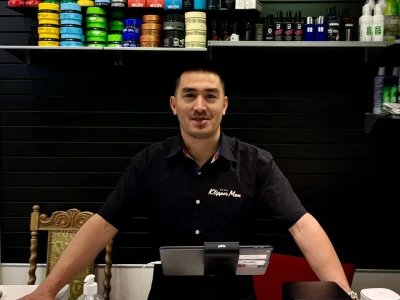 En person står bakom disken i en frisersalong och ler. Bakom dem visar hyllor olika hårstylingprodukter och groomingtillbehör. På disken finns en betalterminal, handsprit och en visitkortshållare.